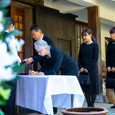 大田区葬儀相談室の受付で高齢の女性が遺族名簿に記帳し列に並ぶ葬儀の様子、黒服で待つ家族に向けた大田区　葬儀の案内